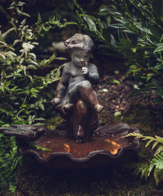 Old birdbath with a sculpture of a sitting child surrounded by tropical plants.
